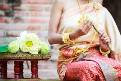 Midsection of young woman in traditional clothing holding flower while sitting against brick wall