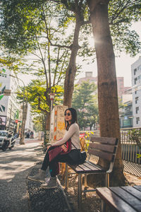 Side view of woman sitting on bench against trees in city
