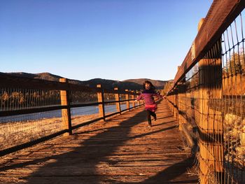 Girl running on footbridge against clear sky