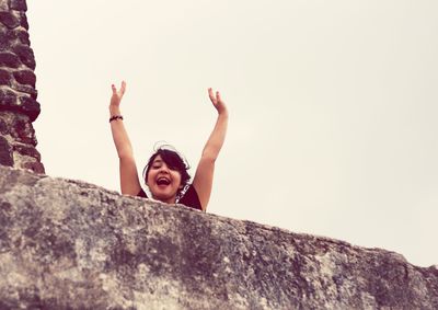 Portrait of smiling woman with arms raised by wall against clear sky