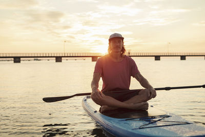 Portrait of man sitting on paddleboard in sea during sunset