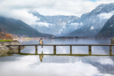 Rear view of woman sitting on bridge over hallstatter see against mountains in hallstatt