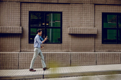 Full length of man holding smartphone walking on footpath