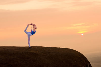 Side view of woman practicing yoga on cliff against orange sky