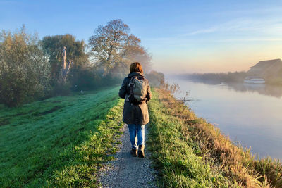 Rear view of a woman walking at sunrise over a green dike along a misty canal