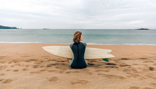 Surfer woman with wetsuit and surfboard sitting on the sand looking aside on the beach