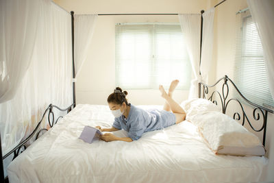High angle view of woman wearing mask using digital tablet lying down on bed at home
