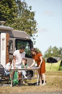 Parents arranging table while siblings sitting against van at trailer park during summer