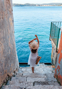 Young woman from behind. woman in white dress standing on stone stairs by sea.