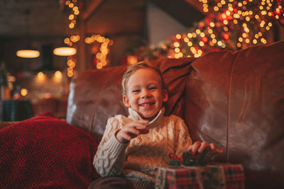 Candid authentic happy child in knitted beige sweater sitting with presents at lodge xmas decorated