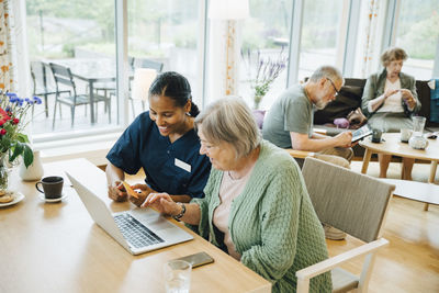 Smiling female healthcare worker assisting senior woman doing online shopping with credit card and laptop in nursing hom
