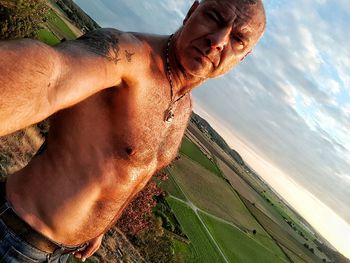 Portrait of serious shirtless man standing on field against cloudy sky