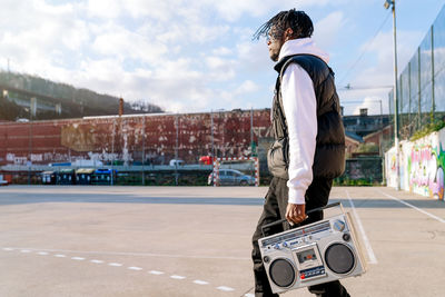 Side view of young african american male in trendy apparel walking on city street with old tape recorder in sunlight