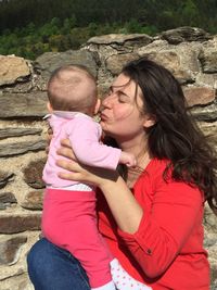 Mother kissing baby while sitting by wall on sunny day