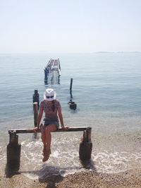 Full length of woman sitting on broken pier at beach against clear sky