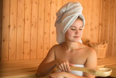 Beautiful young woman holding wooden spoon while sitting in sauna