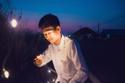Young man holding illuminated string light while sitting on field during dusk