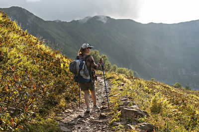 Rear view of young woman with big backpack with trekking pole on mountain trail hiking among plants 
