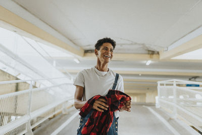 Cheerful young man in bib overalls holding jacket and walking in parking garage