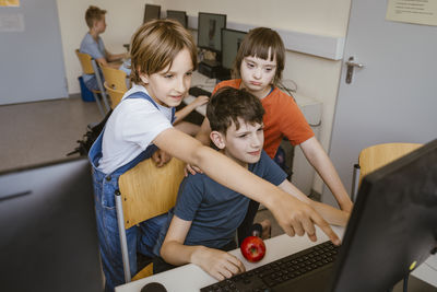 Curious girl pointing at computer near boy and female friend with disability in classroom