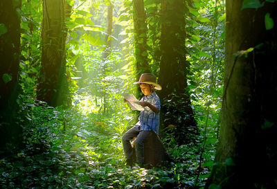 Boy reading map while sitting amidst trees in forest