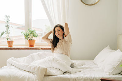 After waking up, the girl stretches while sitting on the bed by the window.