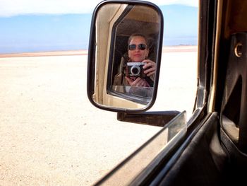 Reflection of woman photographing in side-view mirror of car