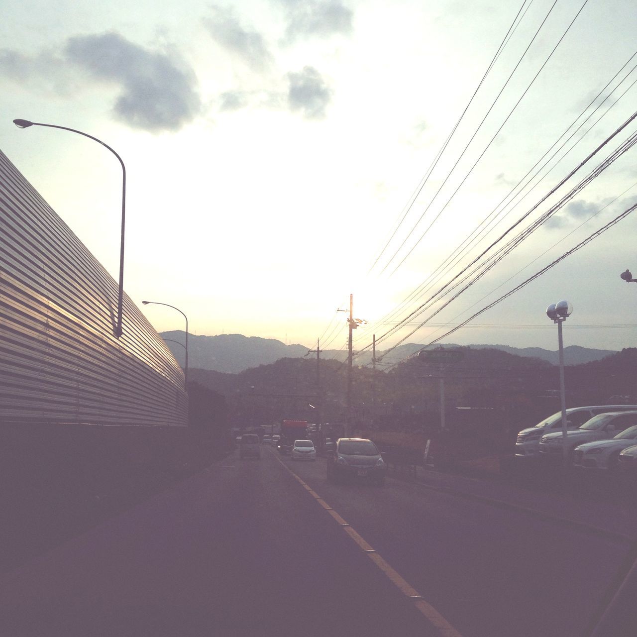 transportation, architecture, car, sky, built structure, building exterior, road, land vehicle, street, city, mode of transport, connection, cable, the way forward, power line, electricity pylon, sunset, street light, cloud - sky, road marking