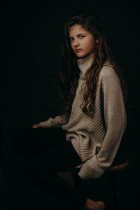 Portrait of cute girl sitting on stool against black background