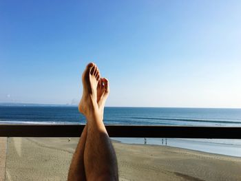 Low section of man relaxing at beach against clear blue sky