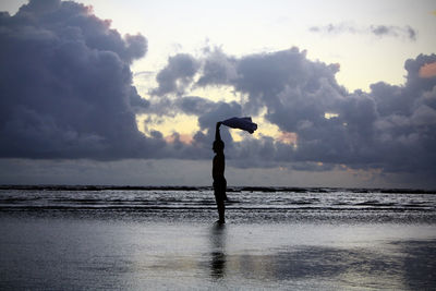 Man holding fabric while standing on shore at beach against cloudy sky