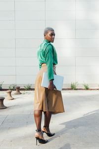 Full length of serious young african american businesswoman with short dyed hair in fashionable outfit standing on street with laptop in hand and looking at camera