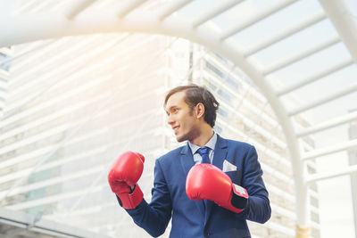 Smiling young businessman wearing red boxing gloves against buildings in city