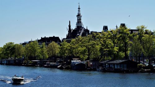 Trees by river against buildings against clear sky