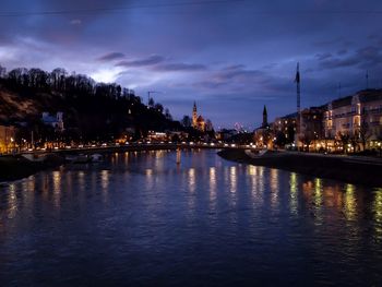 River by illuminated buildings against sky at sunset