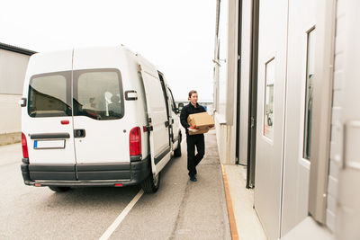 Full length of manual worker carrying cardboard boxes while walking by delivery van