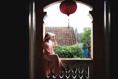 Full length of woman wearing asian style conical hat while sitting on window sill at home