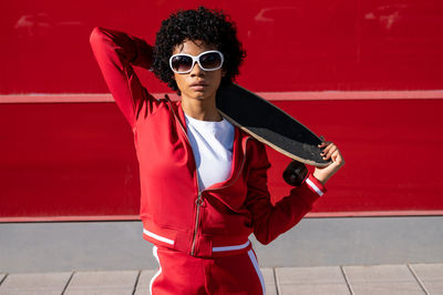 Afro-american woman with short hair and sportswear with a skateboard on red background in city