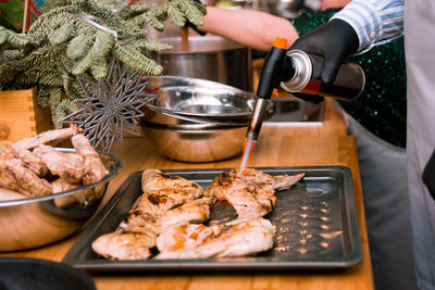 A man's hand holds a burner and roasts chicken meat and chicken wings on a tray