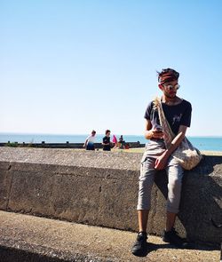 Full length of young man sitting by sea on retaining wall during sunny day