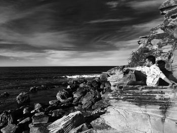 Young man sitting on rock at beach against sky