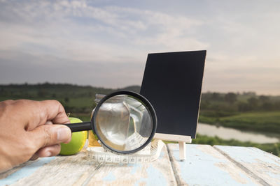 Cropped hand holding magnifying glass at table against sky