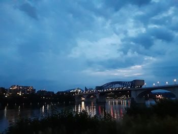 Illuminated bridge over river in city against sky at dusk