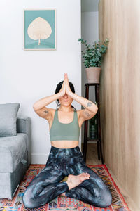 Calm hispanic female in sportswear closing eyes and doing gesturing while sitting in lotus pose on carpet near sofa during yoga session in living room at home