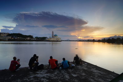 People sitting by lake against sky during sunset