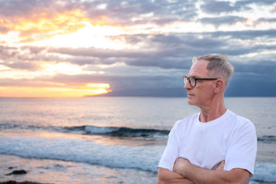 Portrait of young man standing at beach against sky during sunset