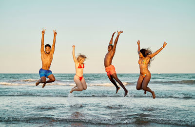 Happy multiethnic men and women raising arms and jumping in sea waves against cloudless sundown sky while having fun on beach in summer evening