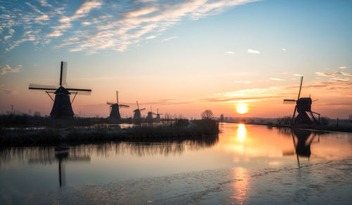 Silhouette windmills by lake against sky during sunset