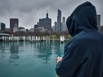 Rear view of man looking across water to buildings against sky in city