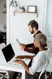 Side view from above of contemporary african american woman sitting at table and working with laptop with young man surfing digital table at home